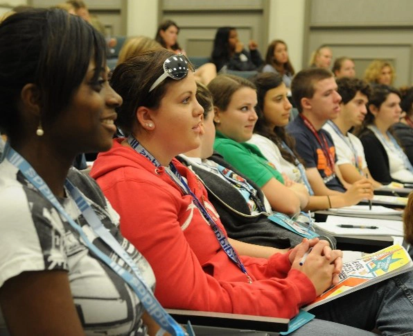 A diverse group of students attentively listening during a workshop or presentation, wearing lanyards with badges in a classroom setting.