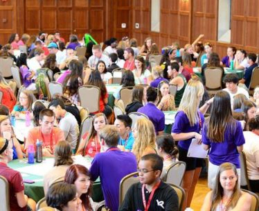 A lively group of participants engaged in discussions at round tables during a conference or workshop in a large event space.