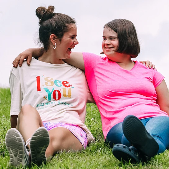 Two female Best Buddies participants embrace while smiling, and sitting in a grassy field.