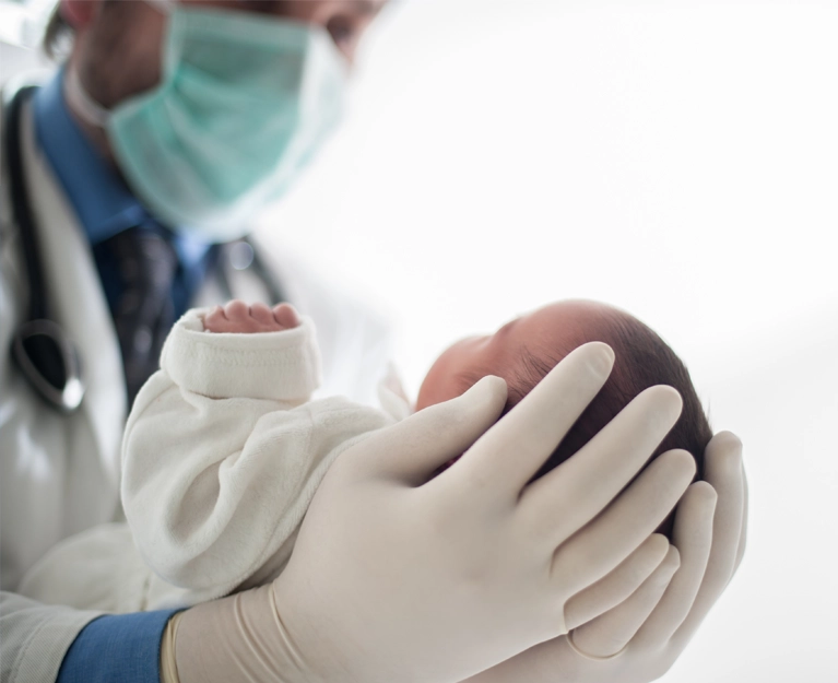 A doctor is holding a baby wearing a surgical mask and gloves.