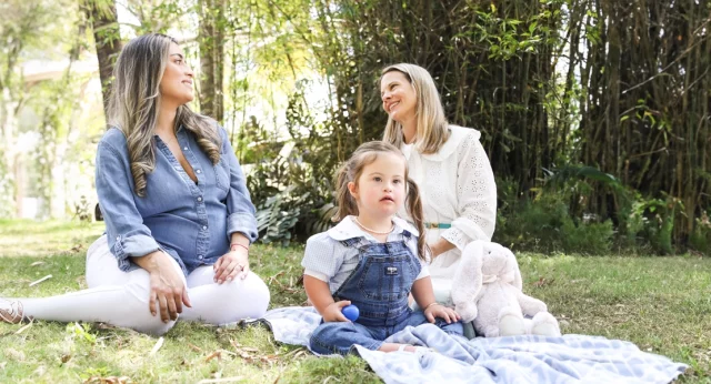 Two women sit on the grass, engaged in a pleasant conversation, with a young girl with Down syndrome sitting between them, playing with a blue toy. They are enjoying a serene moment outdoors, surrounded by lush greenery.