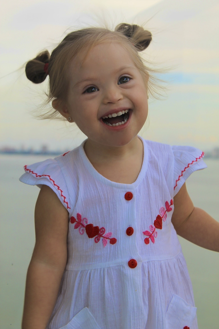 A little girl with Down syndrome smiles brightly, her pigtails framing her face.