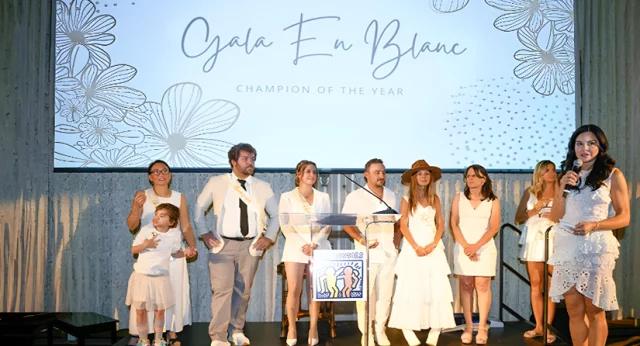 Participants on stage at the Best Buddies International Gala En Blanc, part of the Champion of the Year fundraiser. The group, dressed in white, stands in front of a large screen displaying the event name. The scene reflects the celebratory and formal nature of the event, highlighting the organization's efforts to support individuals with intellectual and developmental disabilities.