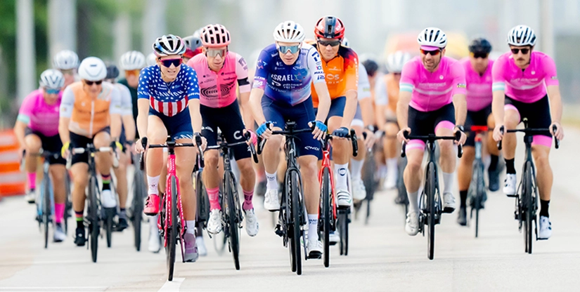 Group of cyclists participating in the Best Buddies Challenge Miami, with riders in various colorful jerseys, including stars and stripes, cycling on a road. Event chaired by Kenneth C. Griffin Charitable Fund and presented by Andrew P. Barowsky Foundation and Mack Cycle.