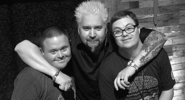 Guy Fieri with his arms around two Best Buddies participants, Chris Gay and Aaron Malman, smiling together in a black and white photo.