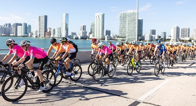 Cyclists riding along a scenic waterfront with a city skyline in the background during the Best Buddies Ride and Gala event.