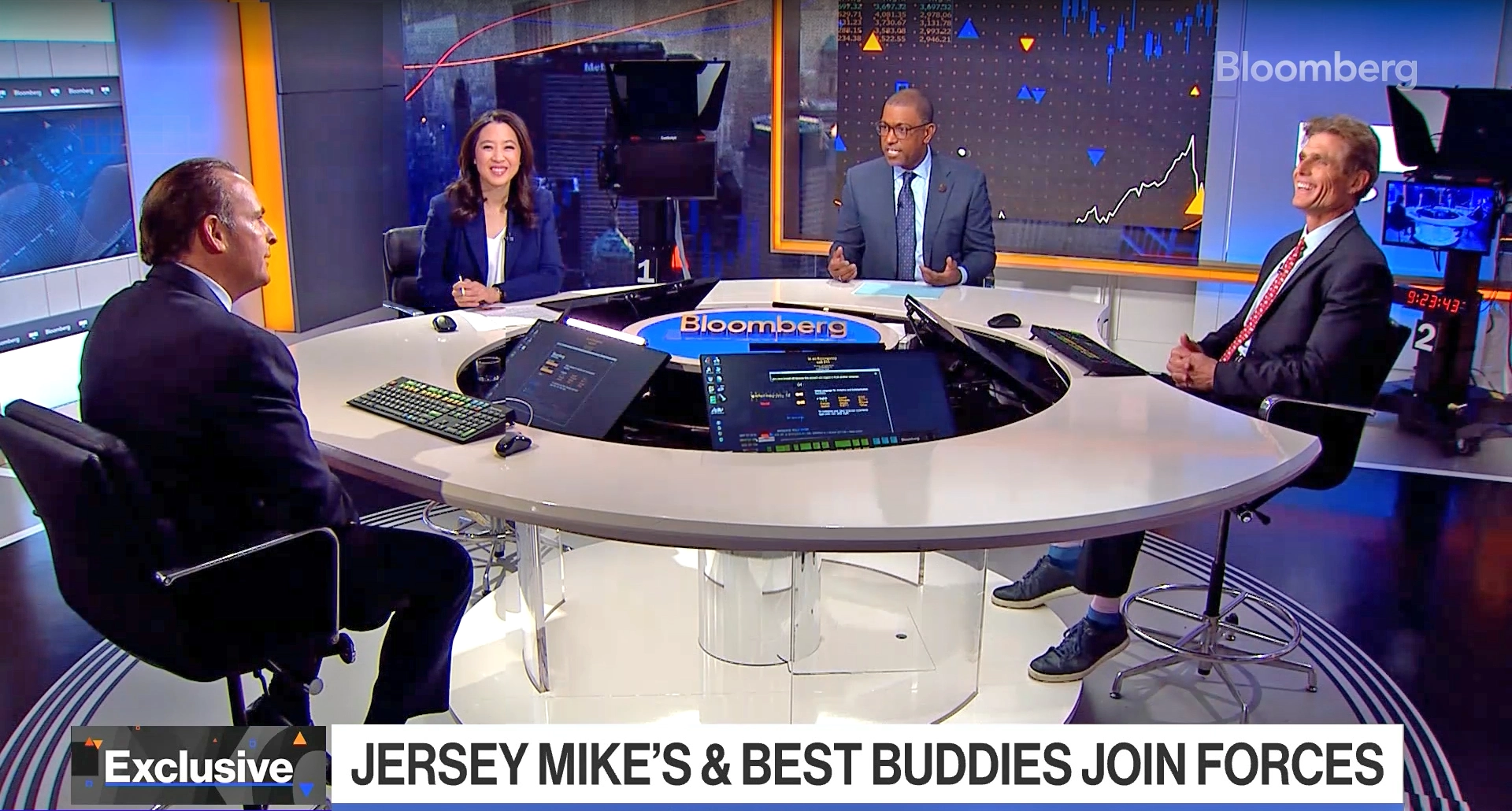 Bloomberg studio discussion featuring four professionals seated around a modern circular desk, discussing the collaboration between Jersey Mike's and Best Buddies, with financial charts displayed in the background.