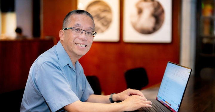 A middle-aged man wearing glasses and a light blue shirt is seated at a table working on a laptop. He is smiling and looking at the camera. The background features a warmly lit space with framed artwork on the wall, creating a professional yet inviting atmosphere.