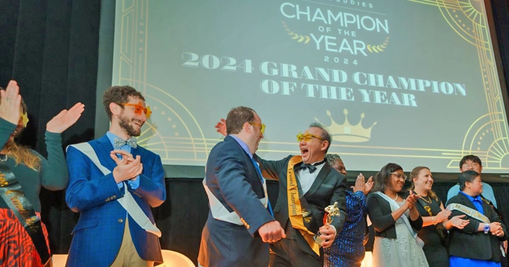 A group of people celebrate on stage at the 'Best Buddies Champion of the Year 2024' event. The backdrop displays '2024 Grand Champion of the Year.' Two men in the center, wearing sashes, share a joyous moment as one holds a trophy. Others around them applaud and cheer, creating an atmosphere of excitement and recognition.