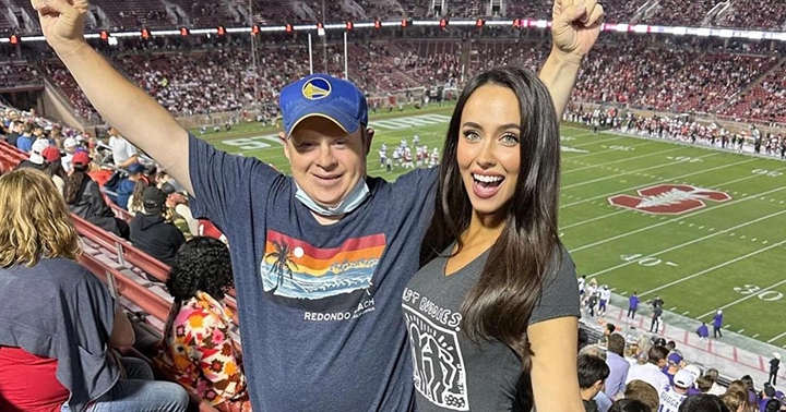 A man and a woman joyfully cheering at a football game in a stadium filled with fans. The man is wearing a blue cap and a Redondo Beach t-shirt, while the woman is wearing a black Best Buddies t-shirt. Both have their arms raised in excitement, celebrating the moment together.