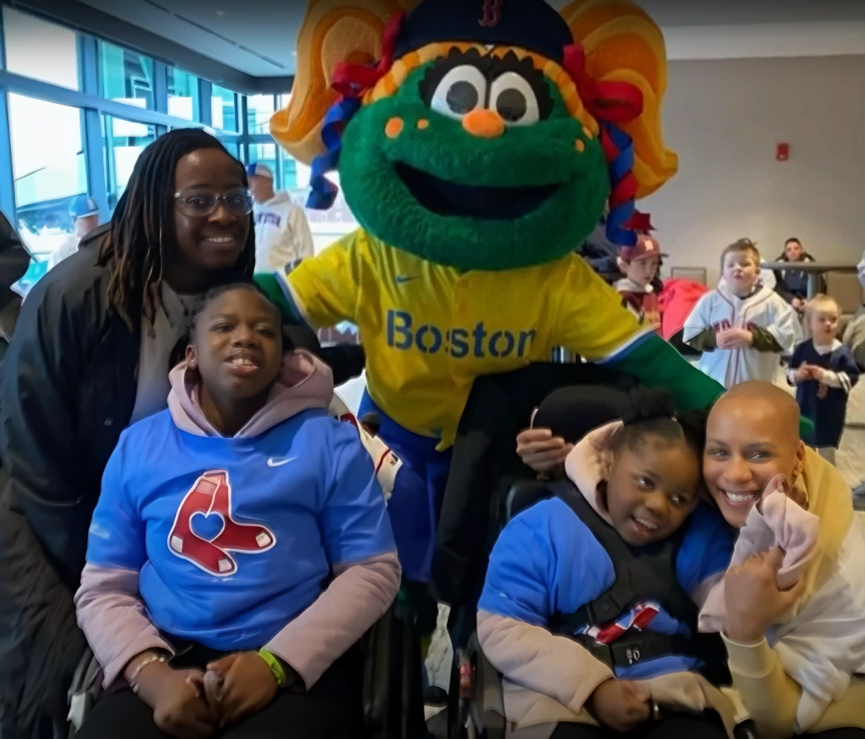 A group photo featuring two children in wheelchairs wearing Boston Red Sox shirts, surrounded by two smiling adults and the Boston Red Sox mascot, Wally the Green Monster, dressed in a yellow Boston jersey, in a cheerful indoor setting with other people in the background.