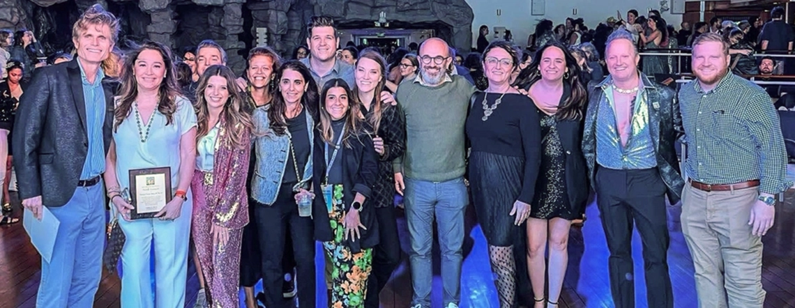 A group of people posing together at a social event, dressed in stylish and festive attire. One individual in the front row holds a plaque, indicating an award or recognition. The background features a lively atmosphere with other attendees and a rock-themed setting.