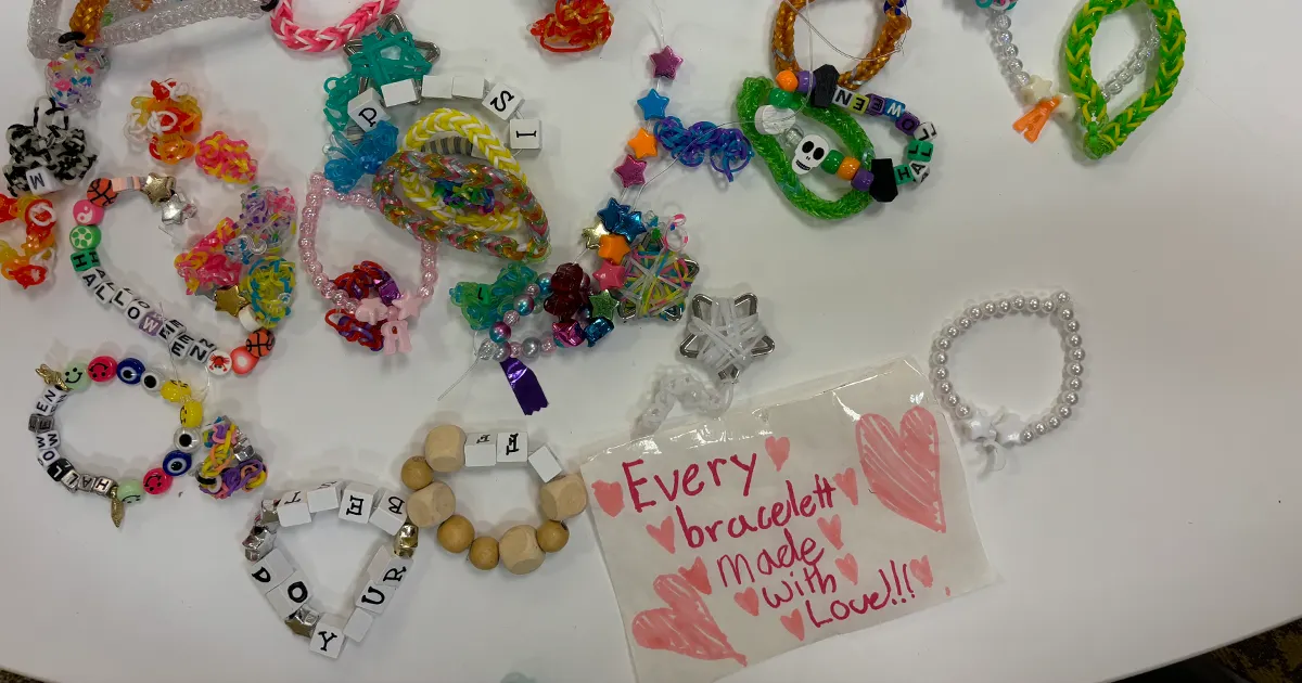 A collection of colorful and unique friendship bracelets displayed on a white table next to a hand-written card with red hearts and the message “Every bracelett [sic] made with Love!!!” in red marker.