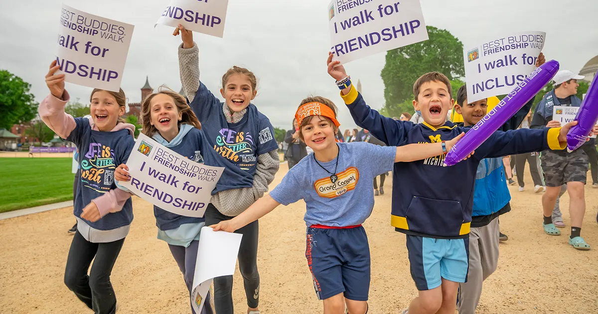 Six elementary school children, including Caroline and Maddie, are running towards the camera and smiling on the National Mall. They are holding signs that say “Best Buddies Friendship Walk. I walk for friendship.”