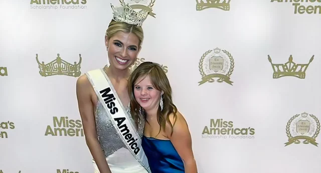 Miss America winner posing with a young woman in a blue dress at a Miss America event, both smiling in front of a branded backdrop.