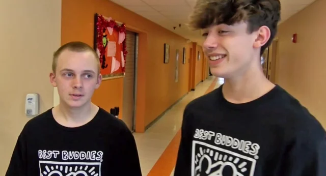 Two students wearing Best Buddies shirts stand in a school hallway, engaging in conversation and smiling.