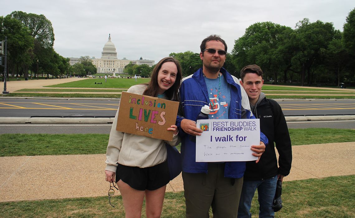 Alex wearing a blue jacket and sunglasses standing in front of the United States Capitol building holding a sign that says “Best Buddies Friendship Walk: I walk for the people that sponsored the walk and the participants. Next to him is Annie, holding a sign that says “inclusion lives here” and Noah. They are all participants of Best Buddies Living Union Market.