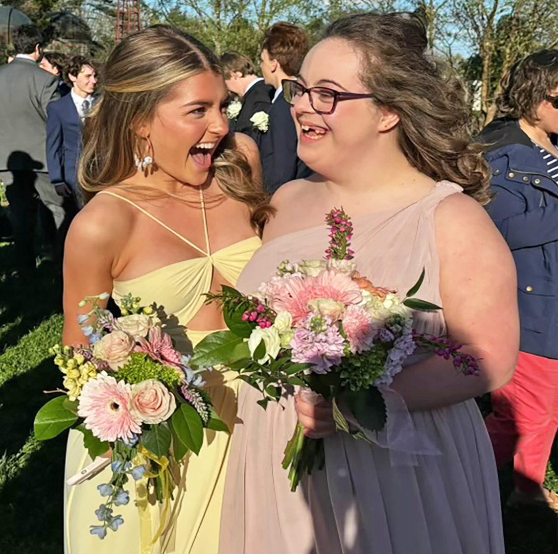 Two young women in elegant gowns share a joyful moment outdoors, holding bouquets of fresh flowers. One wears a yellow dress and reacts with excitement, while the other, in a blush pink gown and glasses, smiles warmly. A crowd in formal attire gathers in the background at what appears to be a celebratory event.