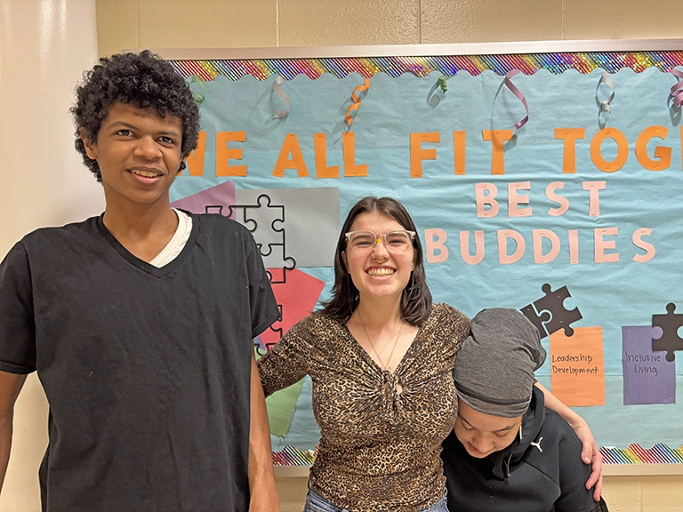 hree friends with their arms around one another smiling in front of a Best Buddies bulletin board.