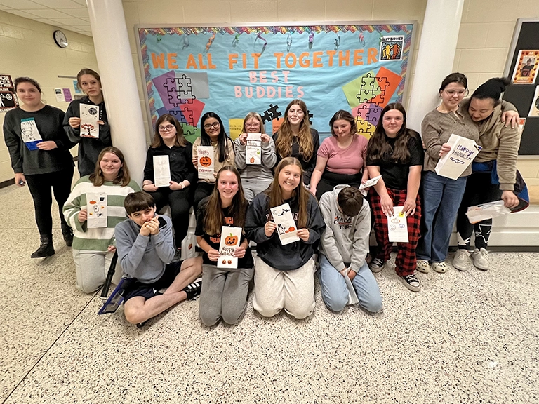 A group of students holding Halloween goodie bags and gathered in front of a Best Buddies bulletin board.