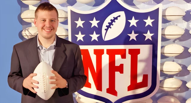 A smiling man in a dark blazer and light blue button-up shirt holds a white football while standing in front of a large NFL logo. The background features multiple white footballs mounted on a blue wall. The setting appears to be an official NFL-related event or display.